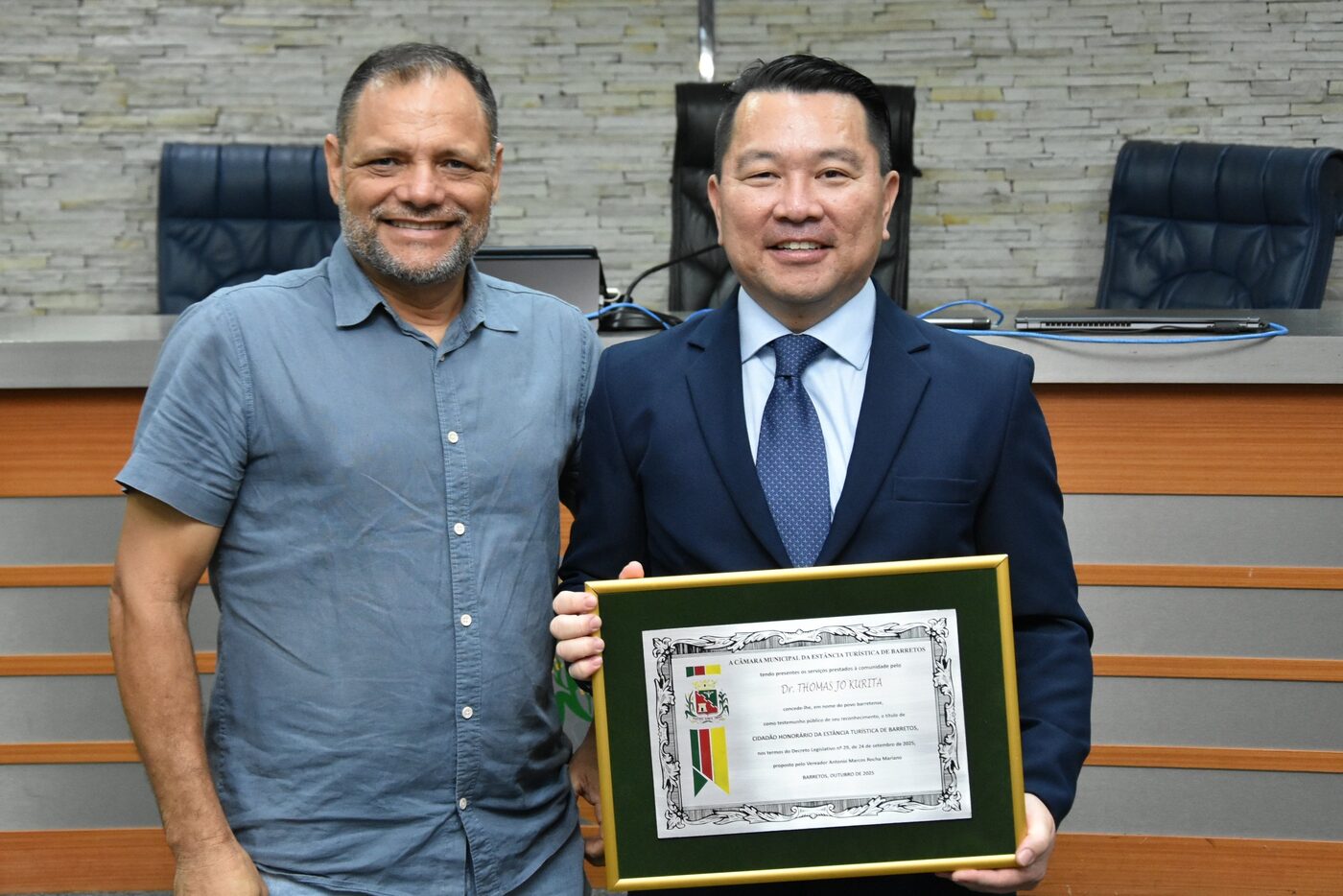 Dr. Thomas Jo Kurita sorri segurando o diploma de Cidadão Honorário, ao lado de outro homem, no plenário da Câmara.