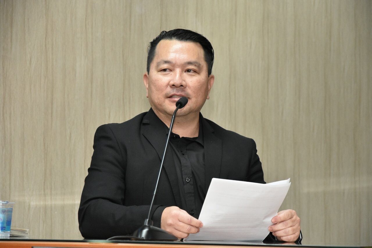 Dr. Thomas Jo Kurita, de terno e camisa pretos, fala ao microfone enquanto segura folhas de papel.